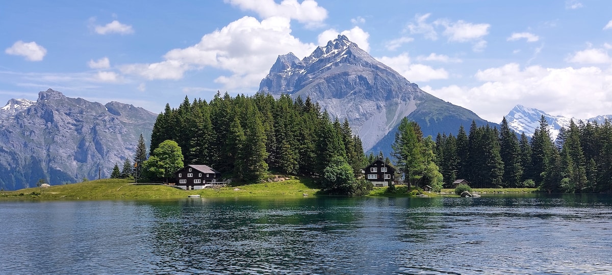 A serene landscape features a calm lake surrounded by lush greenery and towering mountains. Two wooden lodges sit harmoniously among the trees, reflecting the natural beauty of the area. The sky is partly cloudy, adding depth to the stunning mountain backdrop.
