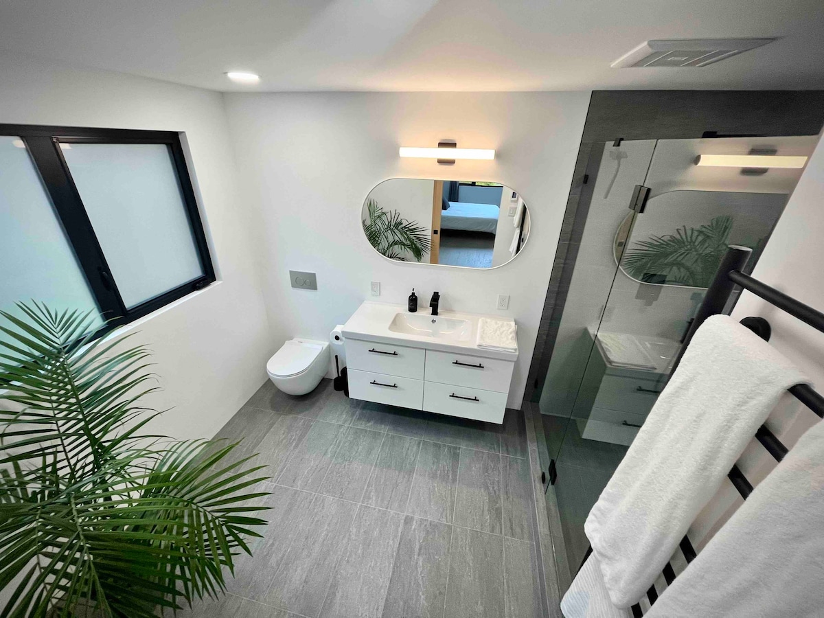 The spacious bathroom features modern fixtures and ample natural light from a window. A sleek vanity with storage is equipped with bottles and a circular mirror. The large shower showcases clear glass walls, while a potted plant adds a touch of greenery to the design.