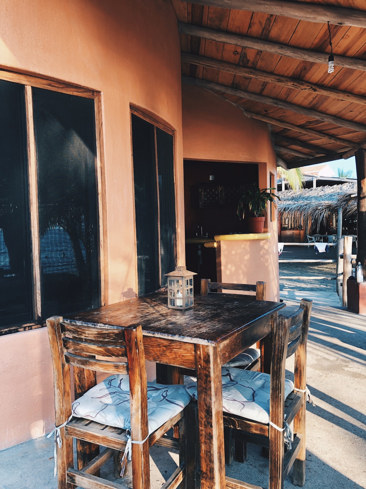 A rustic wooden table with matching chairs is featured on a shaded patio area. The table is adorned with a small lantern, and cushions are placed on the chairs for added comfort. Natural light filters in through large windows and a thatched roof overhead.