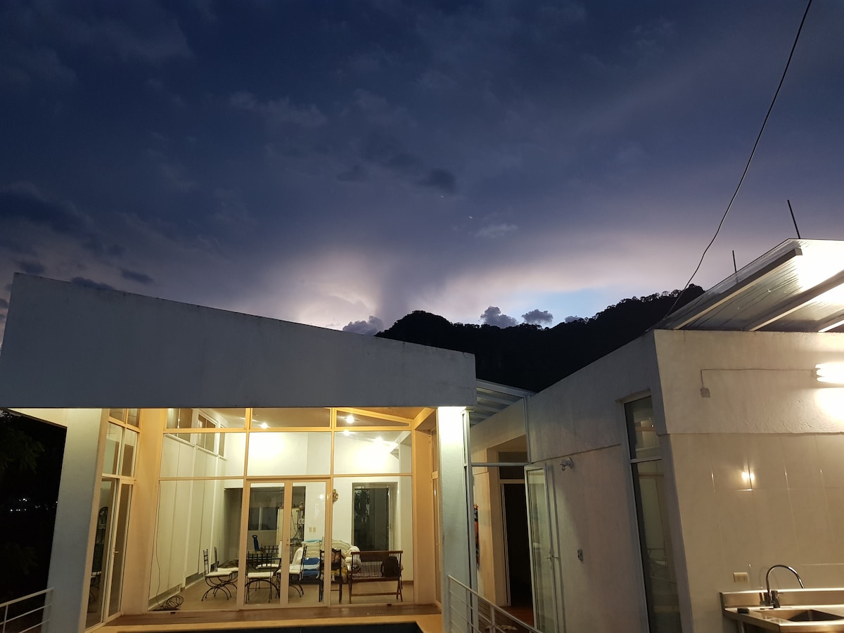The exterior of Casa Shine is bathed in soft twilight, showcasing large glass doors which provide views into the interior space. The silhouettes of surrounding mountains are visible against the evening sky, creating a serene backdrop for the home.