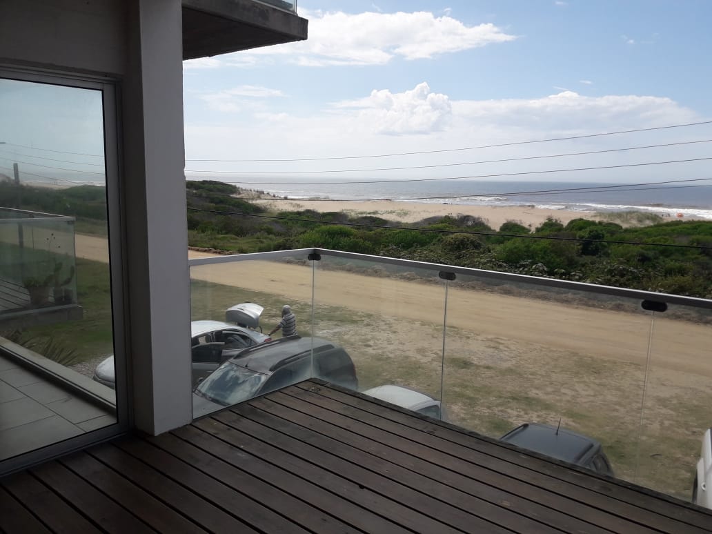 A balcony deck features a transparent glass railing, providing unobstructed views of the beach and ocean. The wooden flooring adds warmth, while the coastal landscape is visible in the distance. Vehicles are parked along the sandy road below.