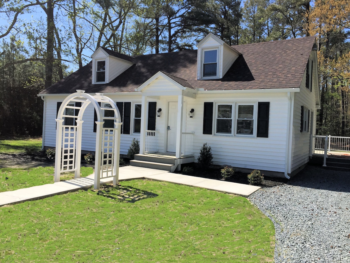A charming white cottage is presented with a sloped roof and landscaped front yard. A wooden archway leads to the entrance, which includes a porch with steps. Several windows provide natural light, and shrubs frame the path to the door.