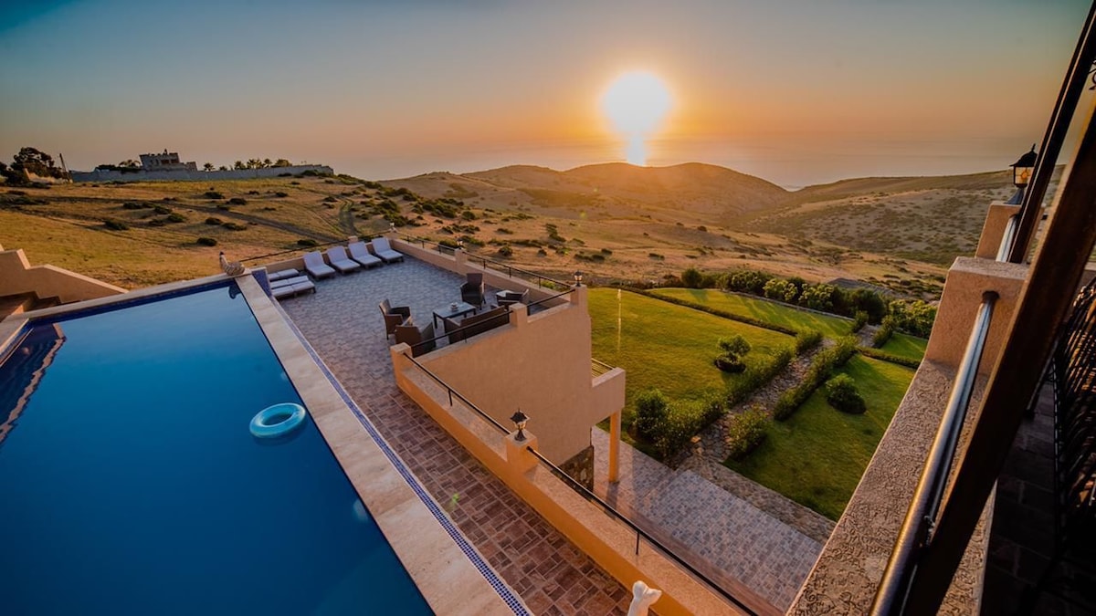 An outdoor terrace features a large swimming pool overlooking expansive hills and the ocean, where the sun sets on the horizon. Lounge chairs are arranged nearby, inviting relaxation while surrounded by greenery and a stone pathway leading through planted areas.