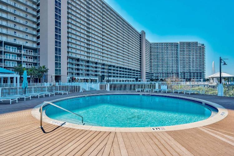 An outdoor pool is showcased with a clear blue water surface, surrounded by a wooden deck. Sun loungers line the perimeter, and tall resort buildings rise in the background, providing a sense of the community atmosphere at Laketown Wharf.