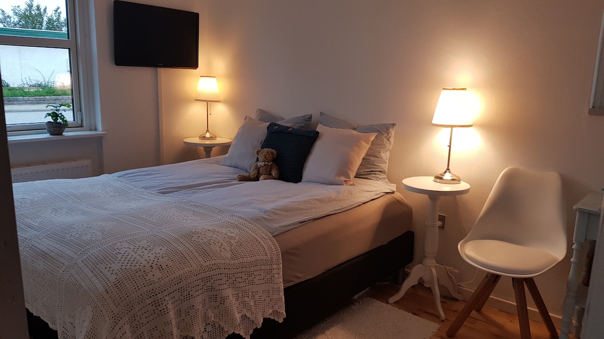 A well-lit bedroom is presented with a spacious bed featuring plush pillows and a lightweight blanket. Two bedside tables support soft-lit lamps, while a wall-mounted television is visible across from the bed. A soft teddy bear rests atop the bedding, adding a touch of comfort.