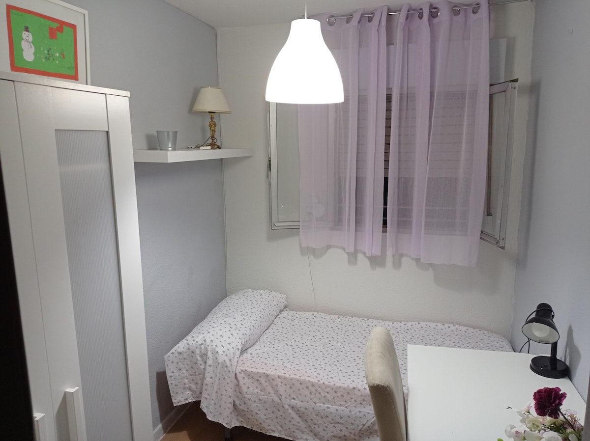 A cozy bedroom features a single bed dressed in white with delicate polka dots. A small desk and chair are positioned beside a window adorned with sheer curtains. A wall-mounted shelf holds decorative items, and a lamp illuminates the space, contributing to a calm atmosphere.