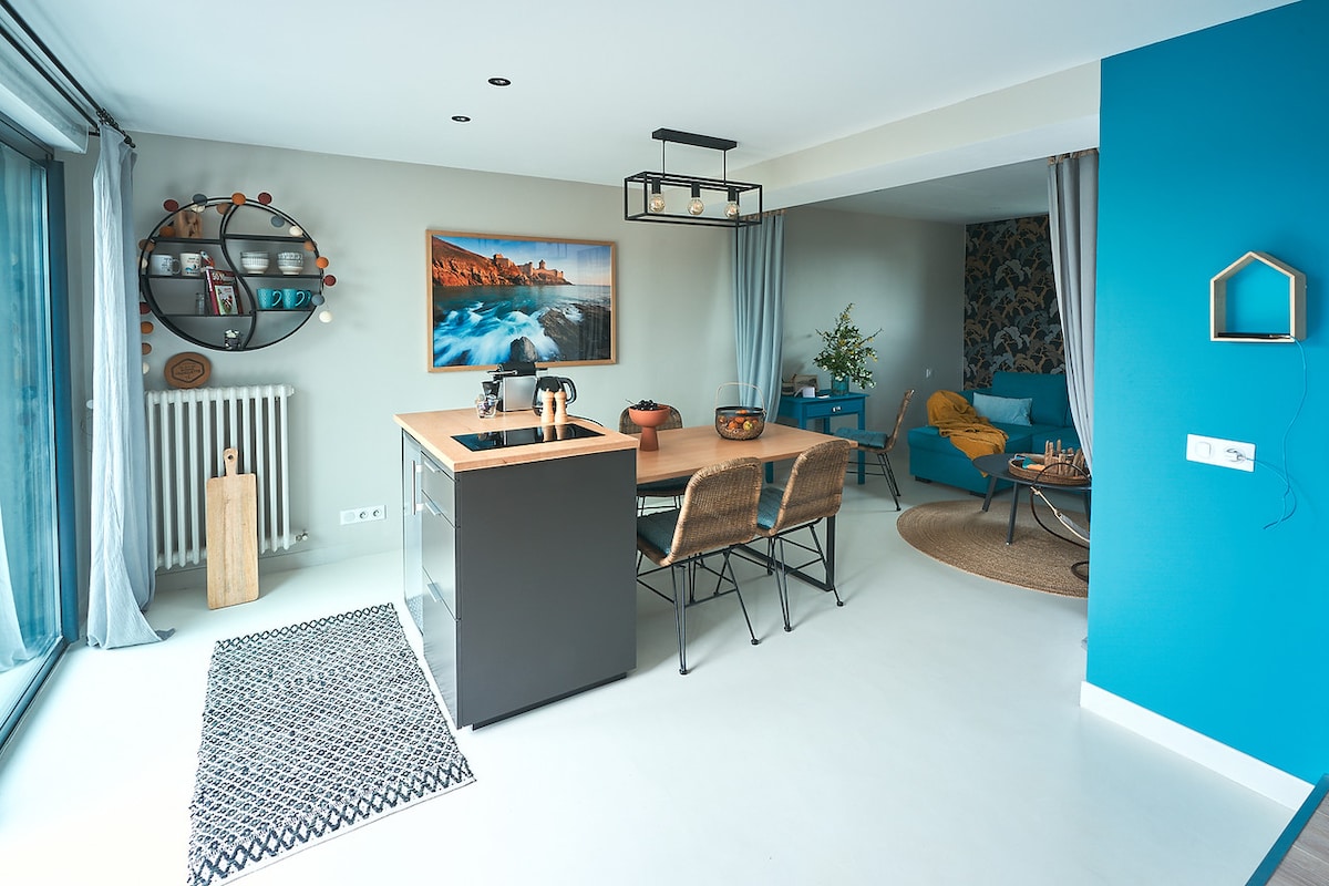 An open-plan living area is presented, featuring a modern kitchen island with bar stools and a stylish dining set. A large window invites natural light, while a vibrant blue wall adds a touch of color. Decorative elements, including wall art and plants, enhance the space.