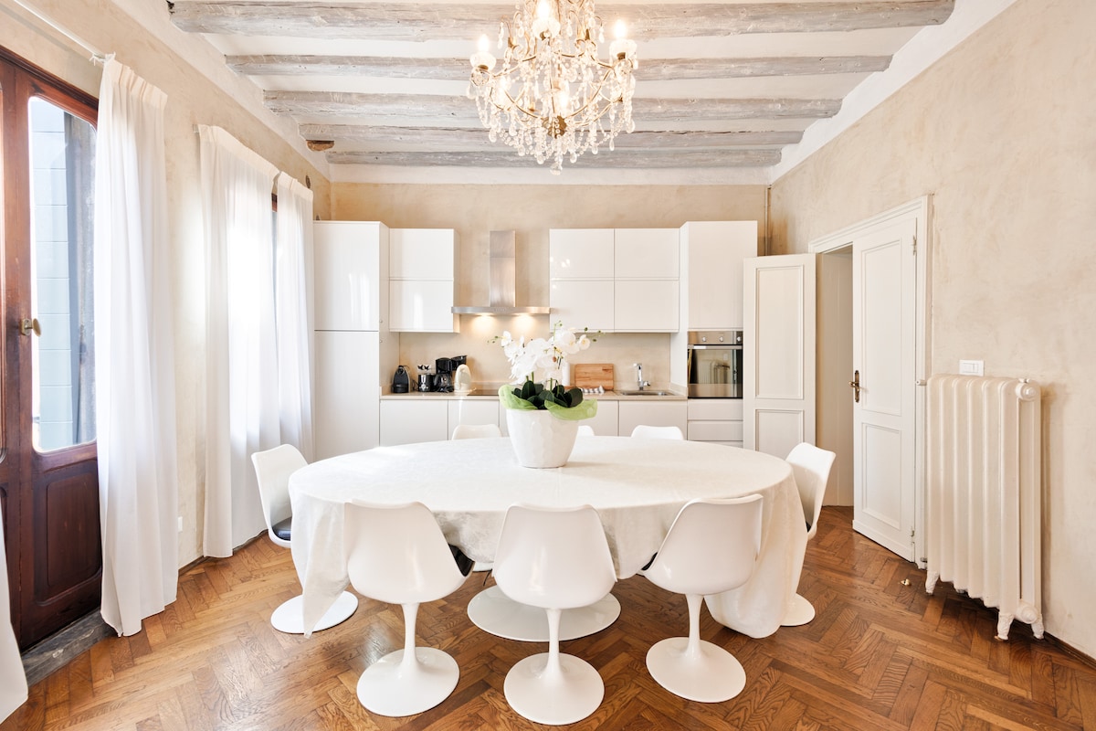 A bright kitchen area features a circular table surrounded by ten white chairs. Natural light floods the space through large windows, highlighting the light-colored cabinetry and wooden beam ceiling. A modern chandelier adds elegance above the table, creating a welcoming atmosphere.