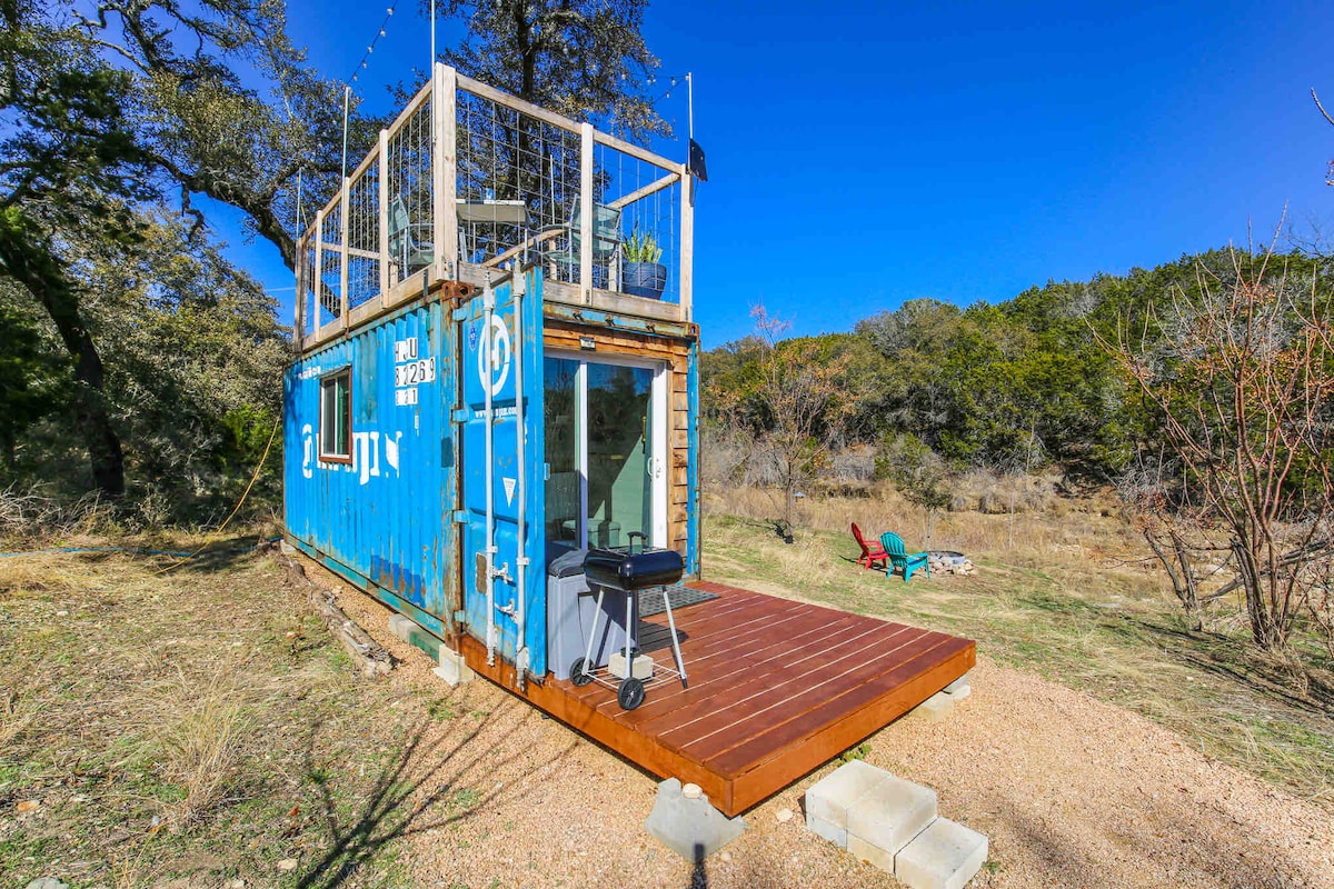A repurposed shipping container home is featured with a wooden deck and grill, offering an outdoor space. The structure is painted blue, surrounded by natural landscape and trees. Comfortable outdoor seating is available nearby, creating an inviting atmosphere for relaxation.