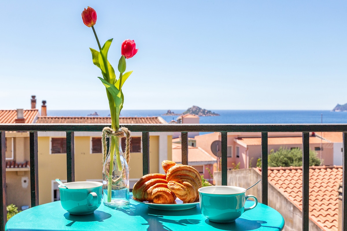 A balcony table is set with two pastel cups and a plate of croissants, accompanied by a vase holding a pair of red tulips. In the background, a clear view of the sea and distant islands is visible beyond a charming neighborhood.