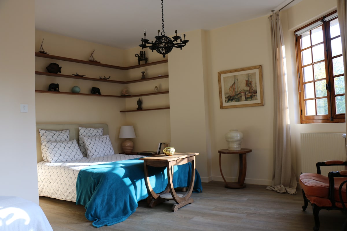 The room features a double bed draped in a soft blue blanket, accompanied by two single beds nearby. A wooden table is centrally positioned, with soft natural light filtering through the window, complemented by shelves displaying decorative items and a framed painting.