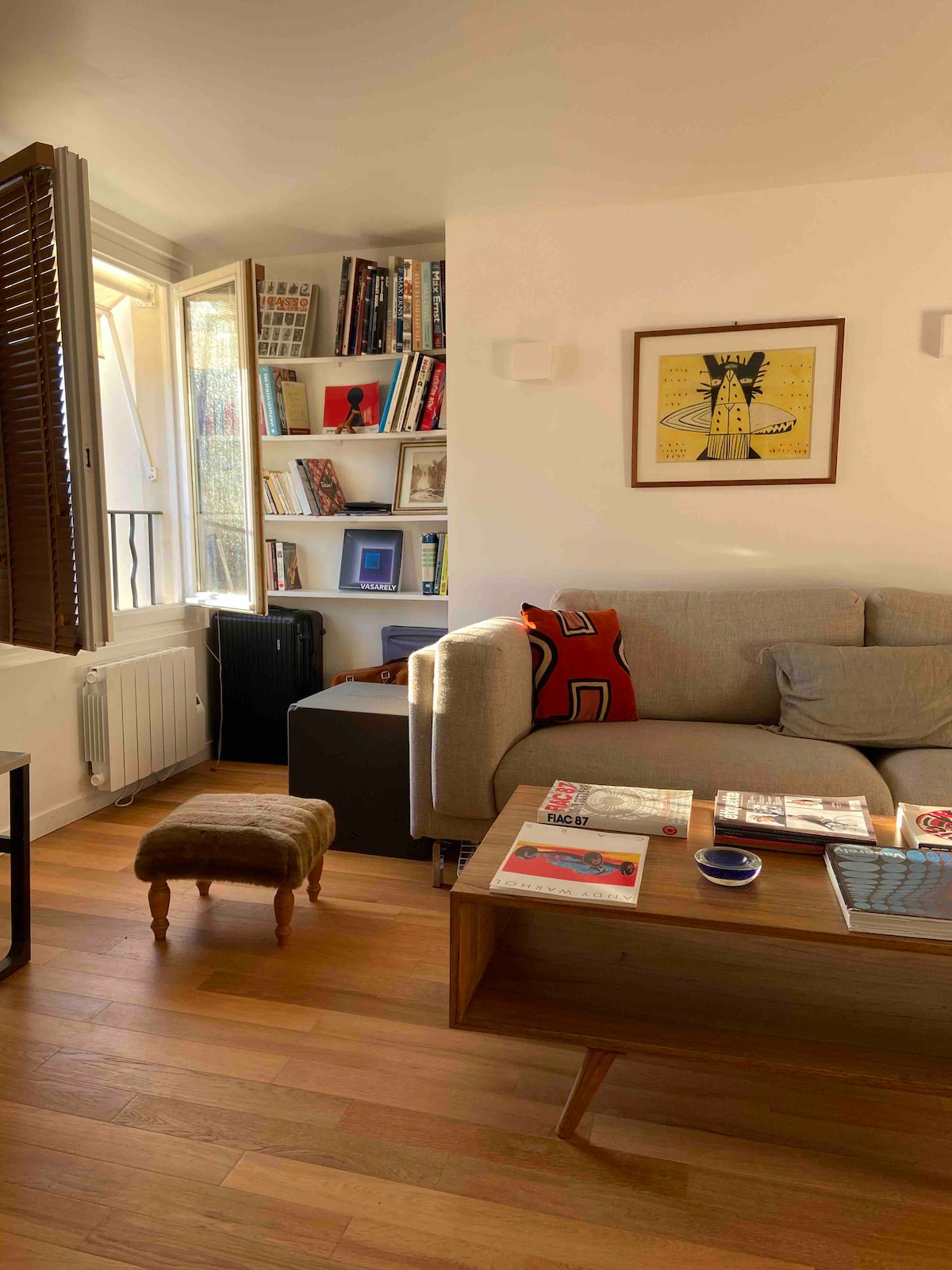 A bright living area features a comfortable sofa adorned with decorative pillows and a colorful throw. A coffee table holds various magazines and a small plate, while a bookshelf filled with books adds character to the space. Natural light enters through a partially opened window.