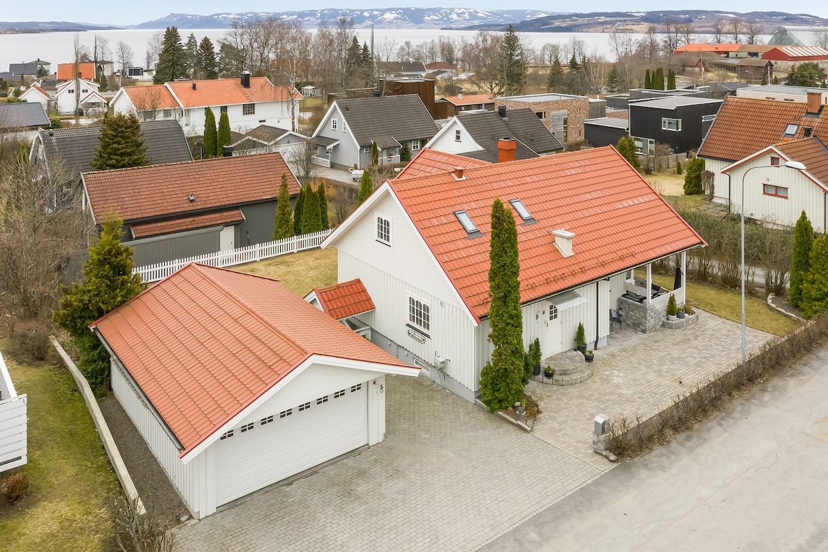 The exterior of a spacious house is visible, featuring a red-tiled roof complemented by green landscaping. A double garage is positioned adjacent to the house, and the surrounding area displays residential properties with views of the distant lake and hills.