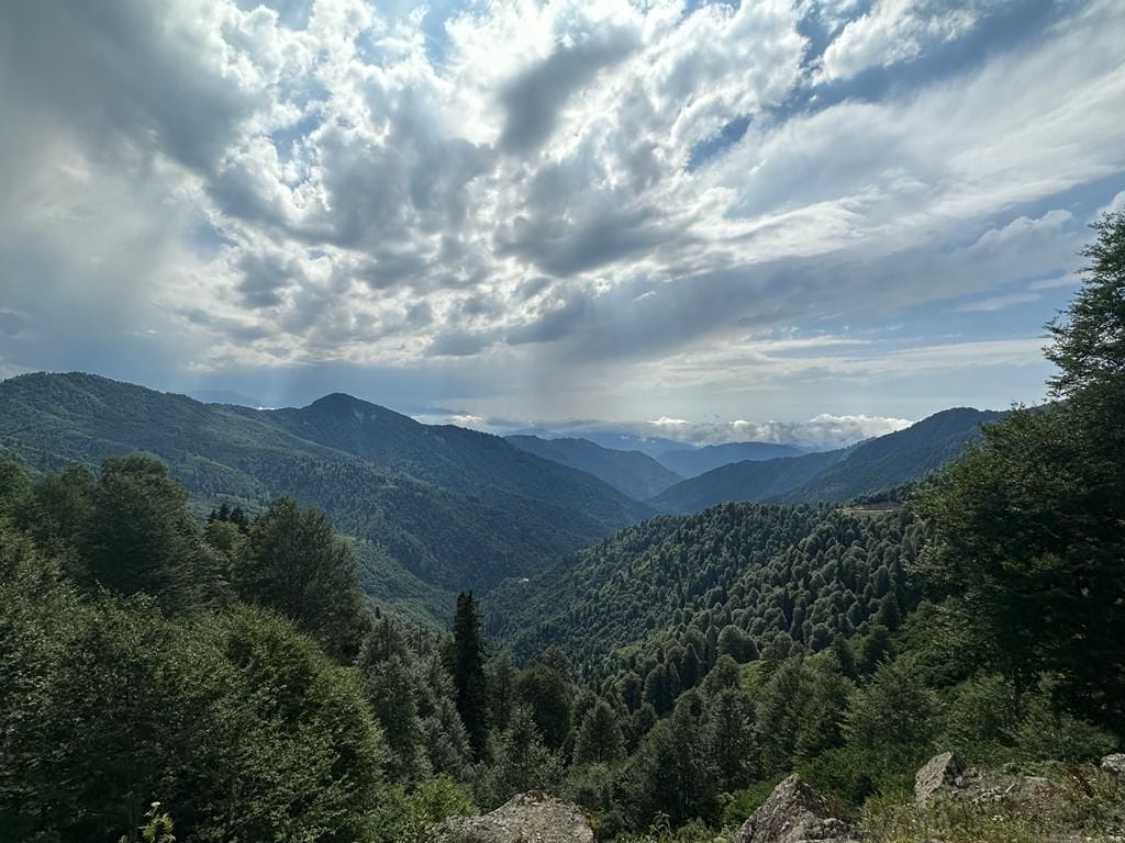 A panoramic view of a lush green mountain landscape is captured under a sky filled with layered clouds. The rolling hills are densely covered with trees, creating a sense of depth and tranquility in the natural environment.
