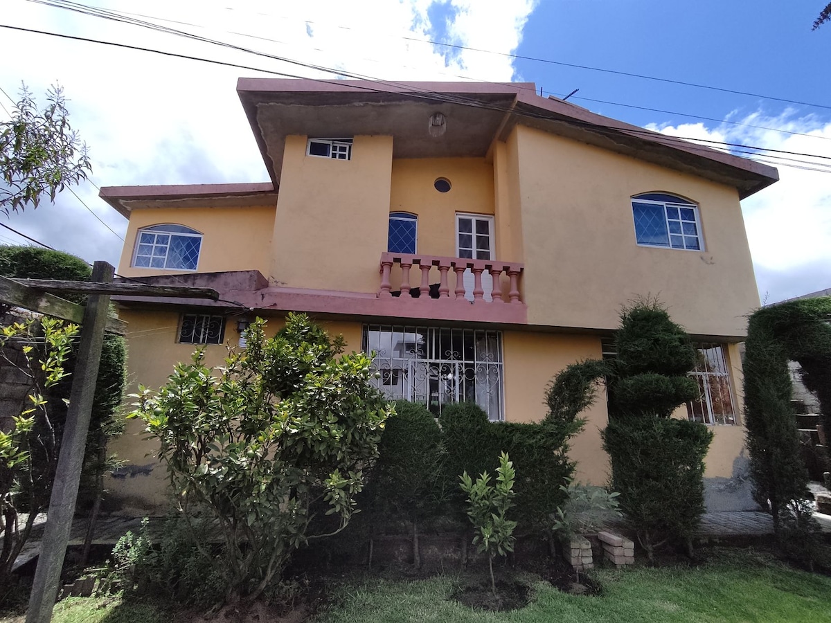The exterior of a two-story house is displayed, featuring a yellow facade and multiple windows that provide natural light. A small balcony is visible on the upper level, while neatly trimmed bushes and trees surround the property, adding greenery to the landscape.