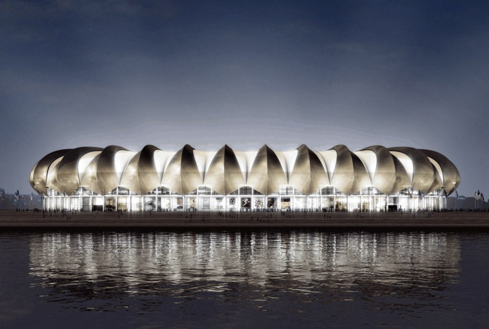 The exterior of a modern stadium is displayed, featuring a unique, wave-like architectural design. The structure is illuminated by bright lights against the backdrop of a nighttime sky, with its reflection visible on the still water in front.