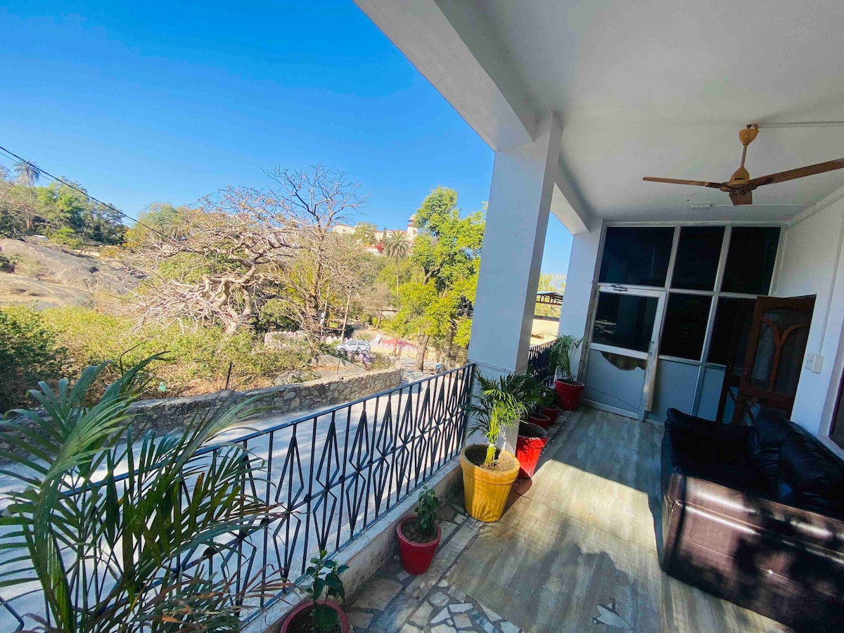 A balcony is featured with a view of surrounding greenery and trees. Potted plants are arranged along the railing, complementing the stone flooring. A ceiling fan hangs above a seating area furnished with dark-colored chairs.