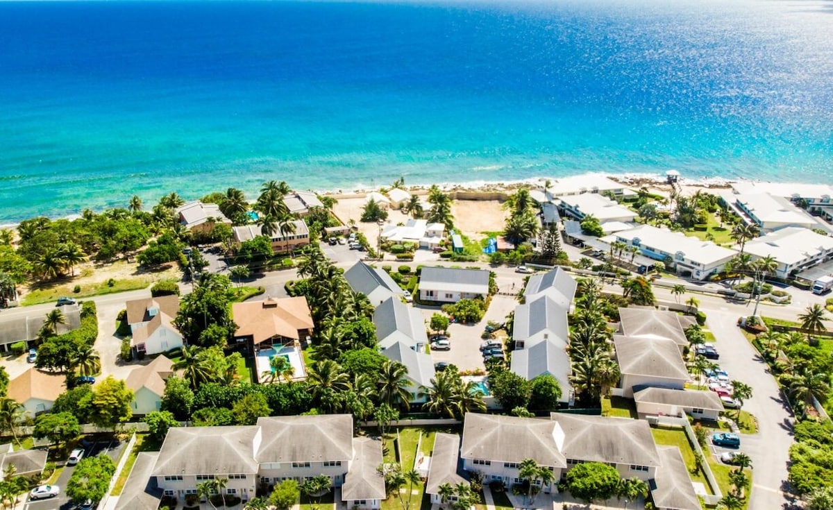 The aerial view captures the vibrant coastline bordered by crystal-clear blue waters. Lush greenery surrounds the residential area, where white-roofed buildings are interspersed among palm trees. The proximity to the beach is highlighted, inviting relaxation and exploration of the nearby waterfront.