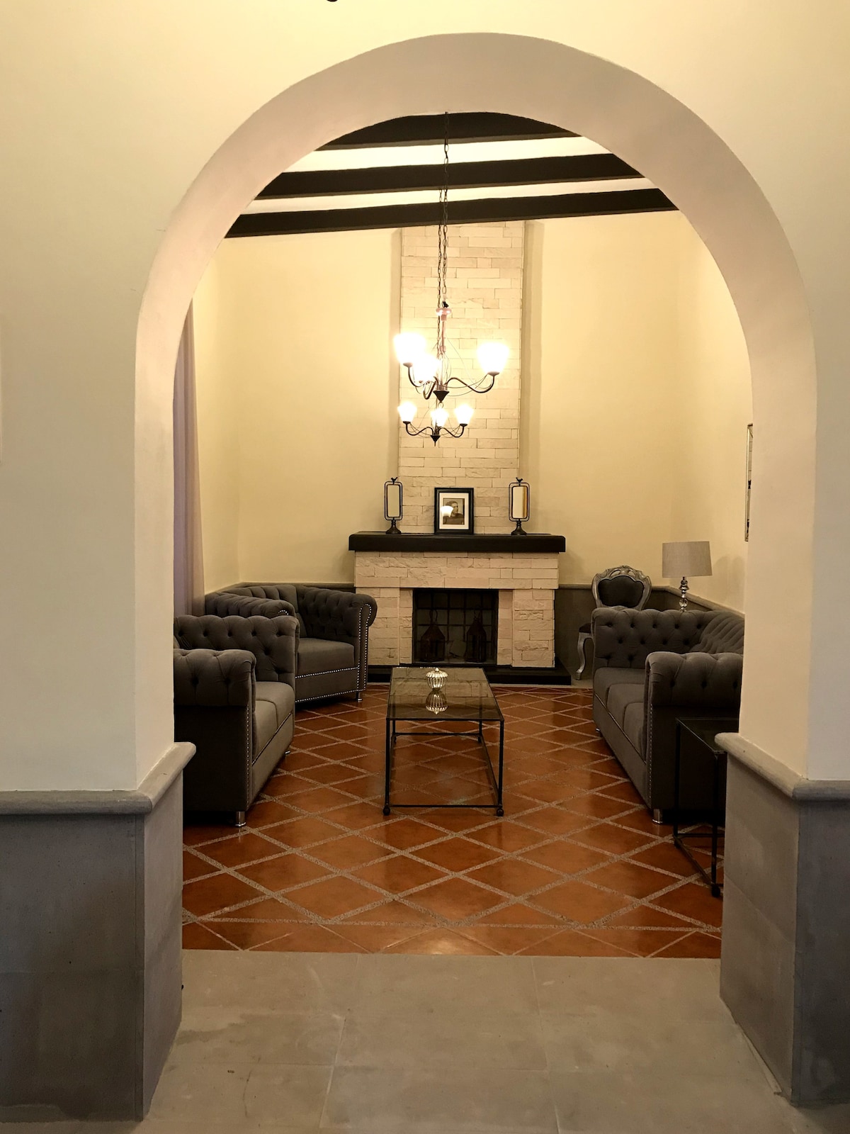 An inviting living space is visible through an arched entrance, featuring two plush tufted sofas flanking a glass coffee table. A fireplace is centered against a light-colored wall, complemented by a chandelier above. Warm tile flooring adds to the cozy atmosphere.