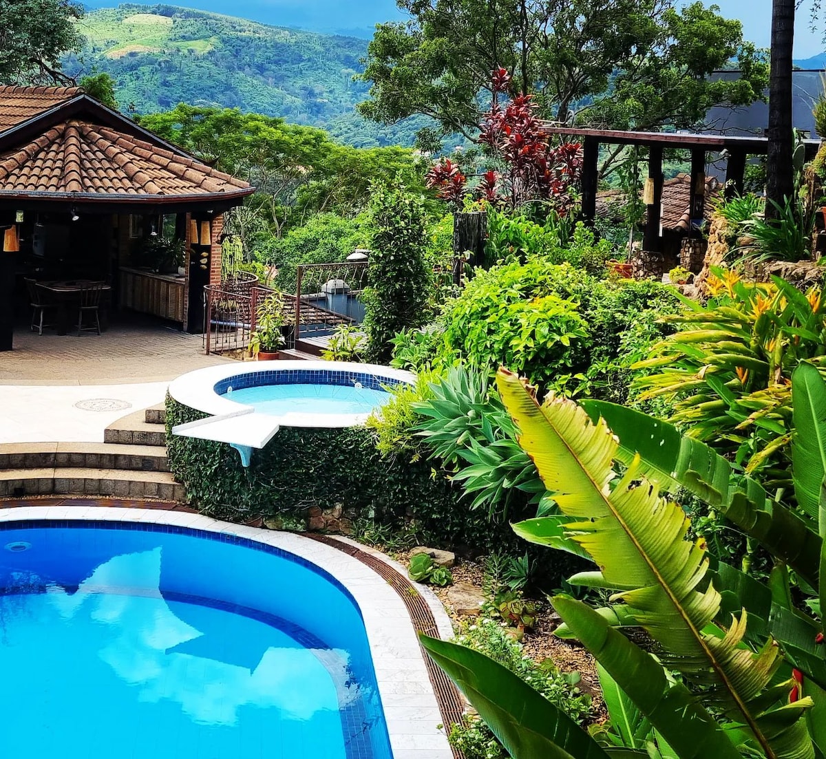 A landscaped outdoor area features a rectangular pool alongside a circular hot tub, both surrounded by lush greenery. A gazebo is visible in the background, enhancing the tranquil environment, while distant mountains create a scenic backdrop.