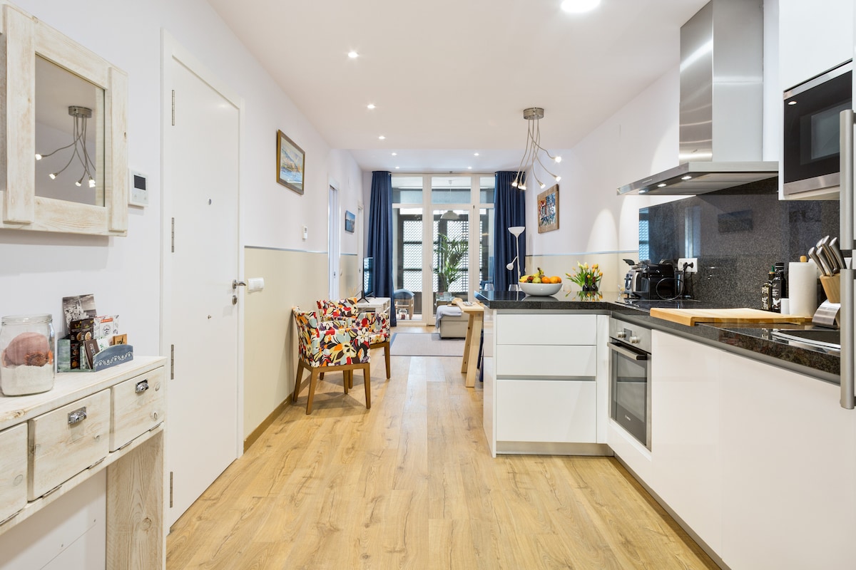 A modern kitchen area is visible, featuring sleek appliances and a wooden countertop. A colorful armchair adds character to the space, while natural light filters through large windows, illuminating the open layout that connects to a cozy living area.