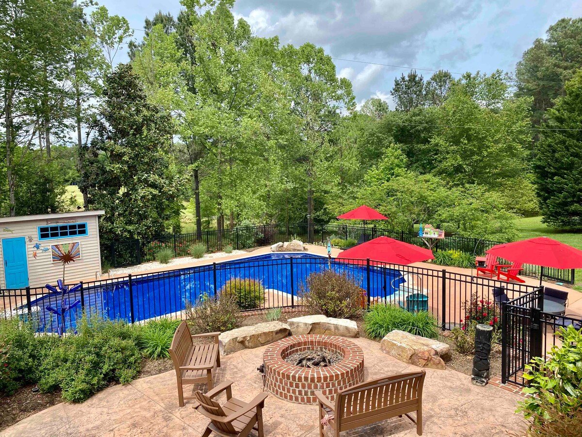 A spacious outdoor area is featured, showcasing a large in-ground pool surrounded by lush greenery. Comfortable lounge chairs and red umbrellas provide shade, while a circular fire pit with wooden seating invites gathering. A fenced perimeter offers safety and privacy in a serene setting.