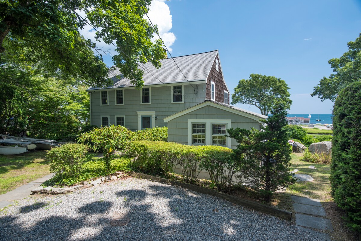 The image shows a charming two-story house with blue siding and a gabled roof, surrounded by lush greenery. A gravel path leads to the entrance, and glimpses of water can be seen in the background, framed by trees and shrubbery.