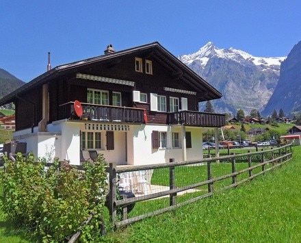 The charming chalet is set against a backdrop of majestic mountains, featuring a wooden exterior and multiple balconies. A private yard enclosed by a wooden fence offers space for relaxation, while the patio is furnished for outdoor seating, inviting guests to enjoy the scenic surroundings.