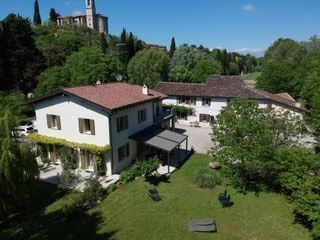 An aerial view showcases the two-story building and adjacent structure surrounded by lush greenery. The area features a spacious garden with shaded seating options, highlighting a serene environment. Nearby trees and a historical landmark are visible in the background.
