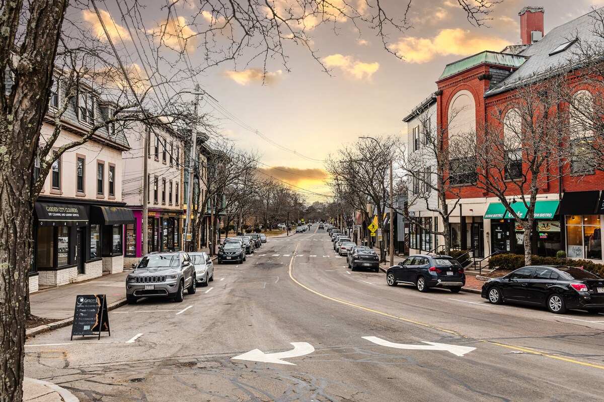 A tree-lined street is viewed from an intersection, showcasing a mix of historic buildings and modern storefronts. Vehicles are parked along the road, and sidewalk cafes are visible. The sky features soft hues of orange and yellow, indicating the time of day is near sunset.