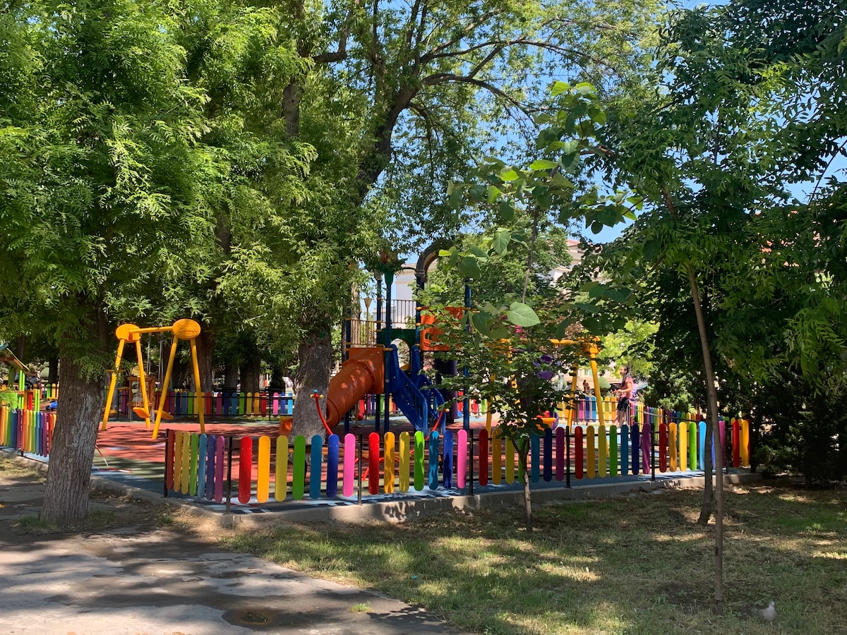 A vibrant playground is surrounded by trees and colorful fencing. Brightly colored swings, climbing structures, and slides offer various play options for children. The shaded area provides a comfortable environment for families enjoying outdoor activities.