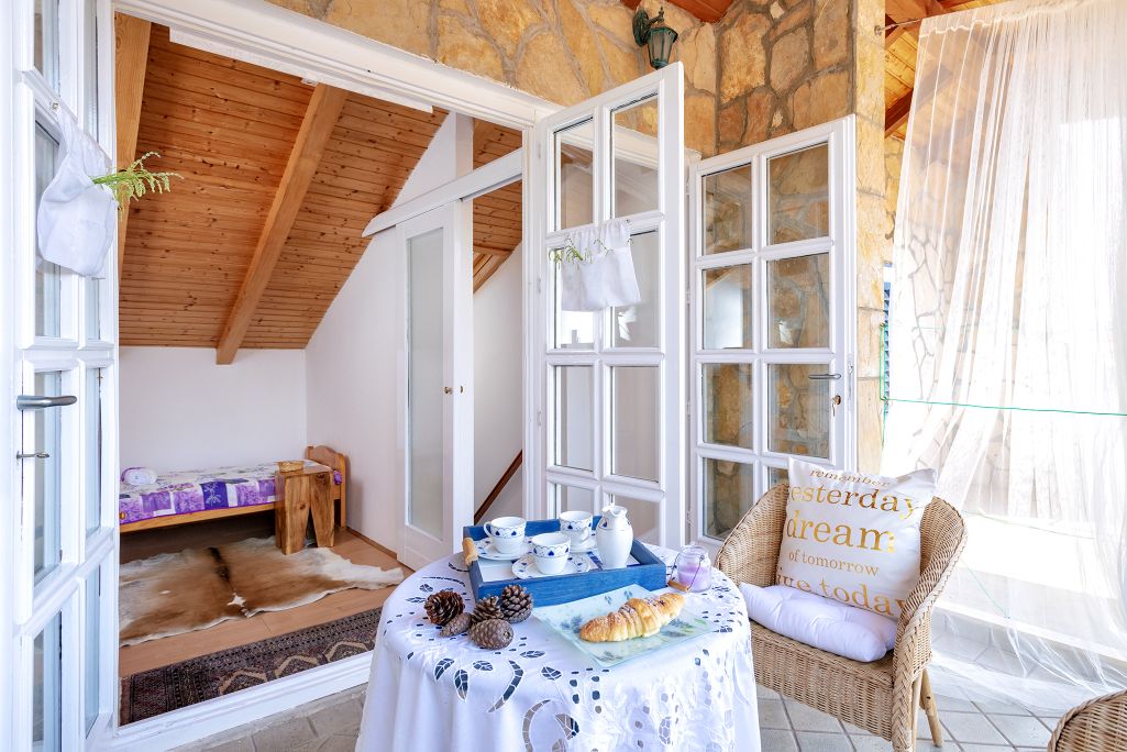 A table set for tea is displayed on a terrace, featuring a white tablecloth and a selection of snacks. Sunlight filters through glass doors, illuminating a cozy sleeping area with wooden beams and a bed in the background. A comfortable chair, adorned with a decorative pillow, invites relaxation.