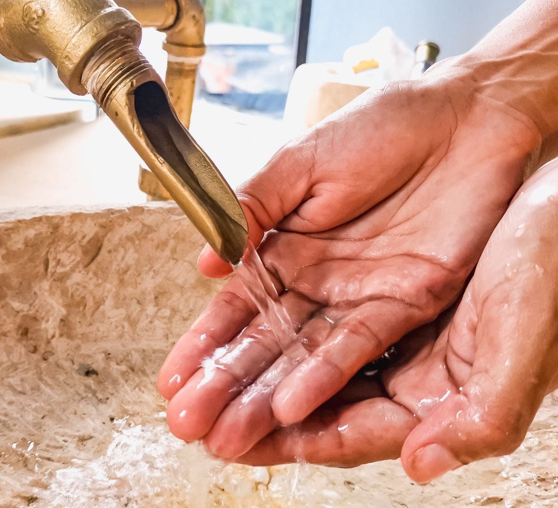 Two hands are positioned under a brass faucet, capturing a gentle flow of water. The sink features a textured stone basin, complementing the warm metallic tones of the faucet. Light reflects off the water, highlighting the details of the hands and the sink.