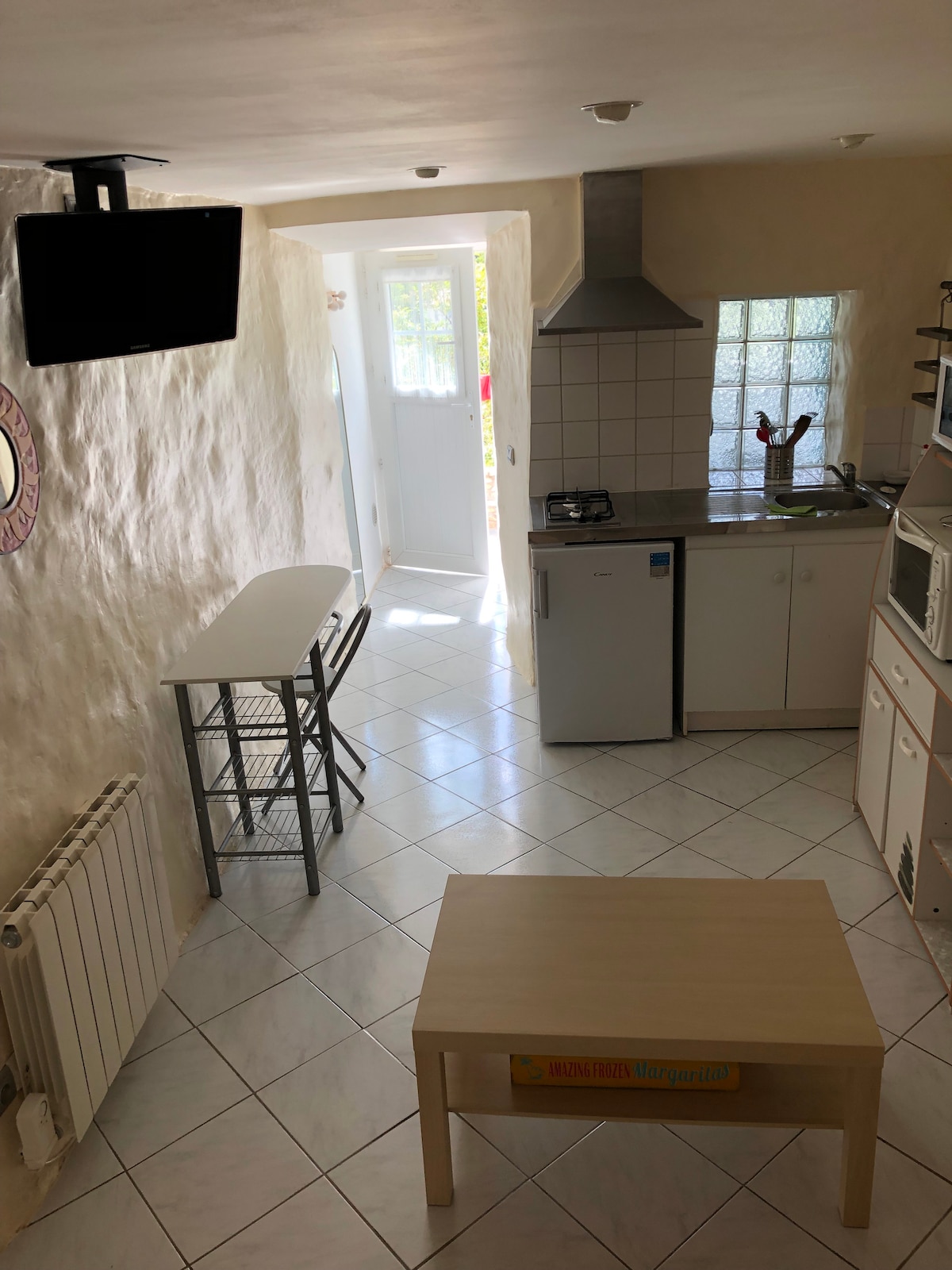 The kitchen area features white walls and tiled flooring. A small table with two high stools is positioned nearby. A modern stove and refrigerator are visible, along with a microwave. Natural light enters through the window near the entrance, which is partly obscured by a wall.