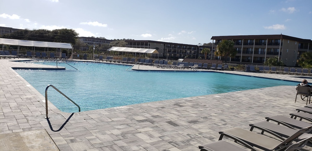 A large pool with a clear blue surface is surrounded by a spacious patio area featuring stone flooring. Lounge chairs are arranged along the edge, with palm trees visible in the background near the multi-story building. Shade structures are positioned at one end of the pool.