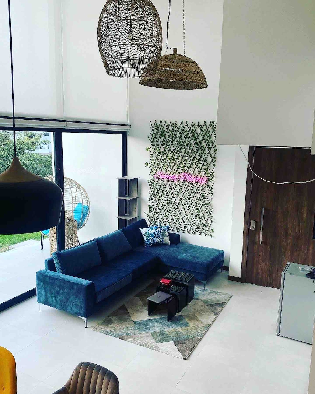 A spacious living area features a modern sectional sofa in blue, positioned against a wall adorned with decorative greenery. A coffee table sits on a patterned rug, while unique light fixtures hang from the high ceiling, enhancing the airy ambiance.