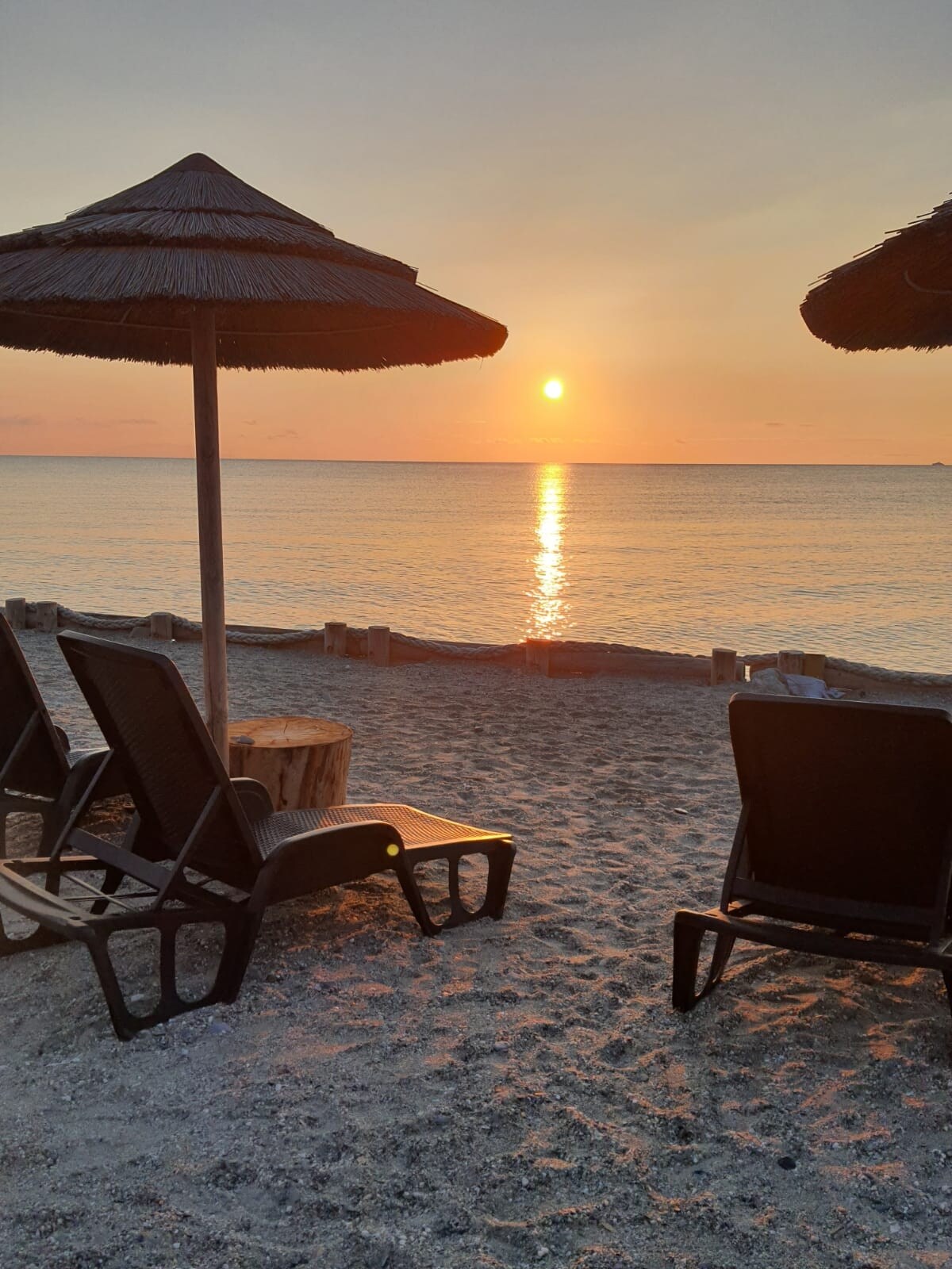 Two lounge chairs are positioned on fine sand, facing the serene sea. A straw umbrella provides shade, while the setting sun casts a warm orange glow across the water. The horizon reflects light, creating a tranquil atmosphere.