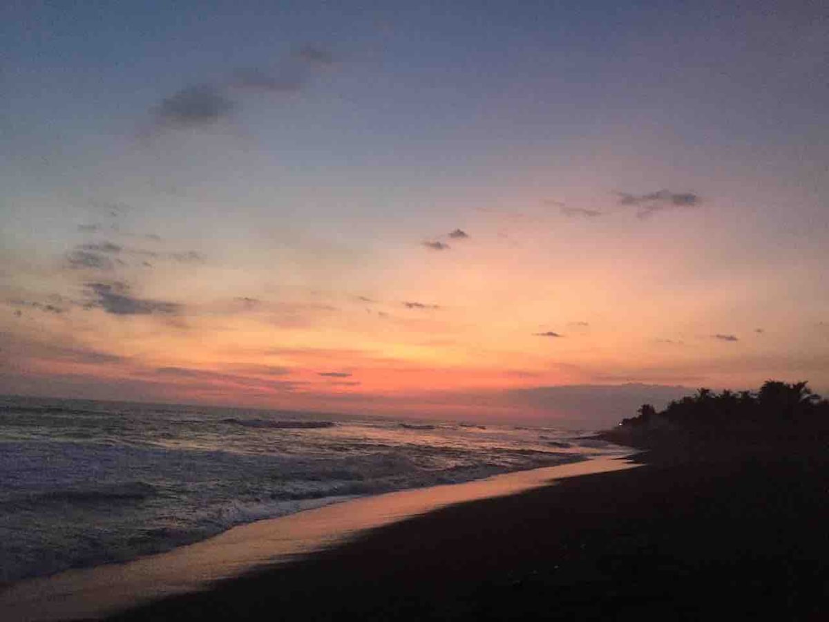 A serene beach scene captured at sunset, featuring gentle waves lapping at the shore. The sky is painted with soft hues of orange, pink, and purple as the sun sets on the horizon. Silhouettes of palm trees are visible in the distance.