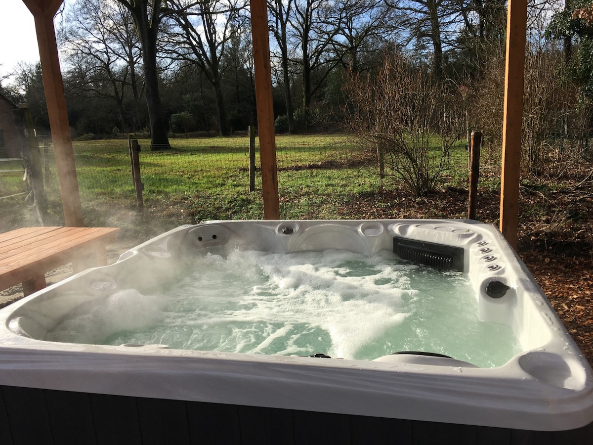 A hot tub with bubbling water is situated under a wooden shelter, surrounded by lush greenery and trees. Gentle steam rises from the surface, creating a serene environment. A wooden table can be seen nearby, complementing the outdoor ambiance.