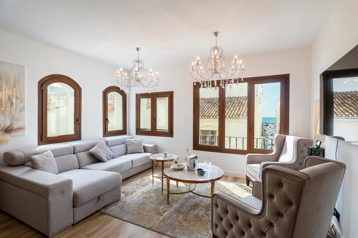 A spacious living room is highlighted by large windows, allowing natural light to fill the area. Two elegant chandeliers provide illumination, while a comfortable sectional sofa and a classic armchair offer seating. A decorative round coffee table sits centrally on a soft area rug.