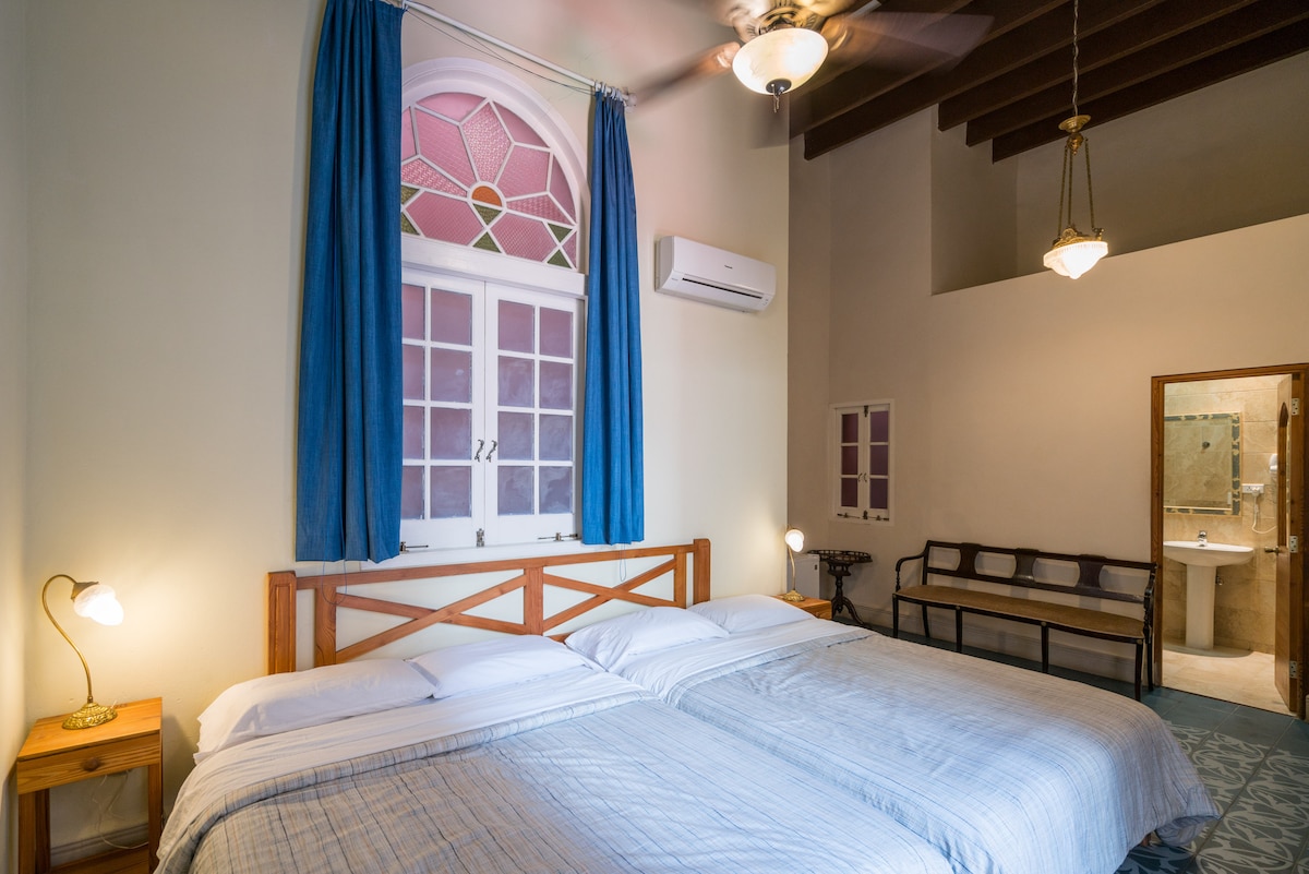The room features two spacious beds dressed in light blue linens, set against a backdrop of high ceilings and a large window adorned with colorful stained glass. Soft lighting is provided by bedside lamps, and a small seating area is visible in the corner.