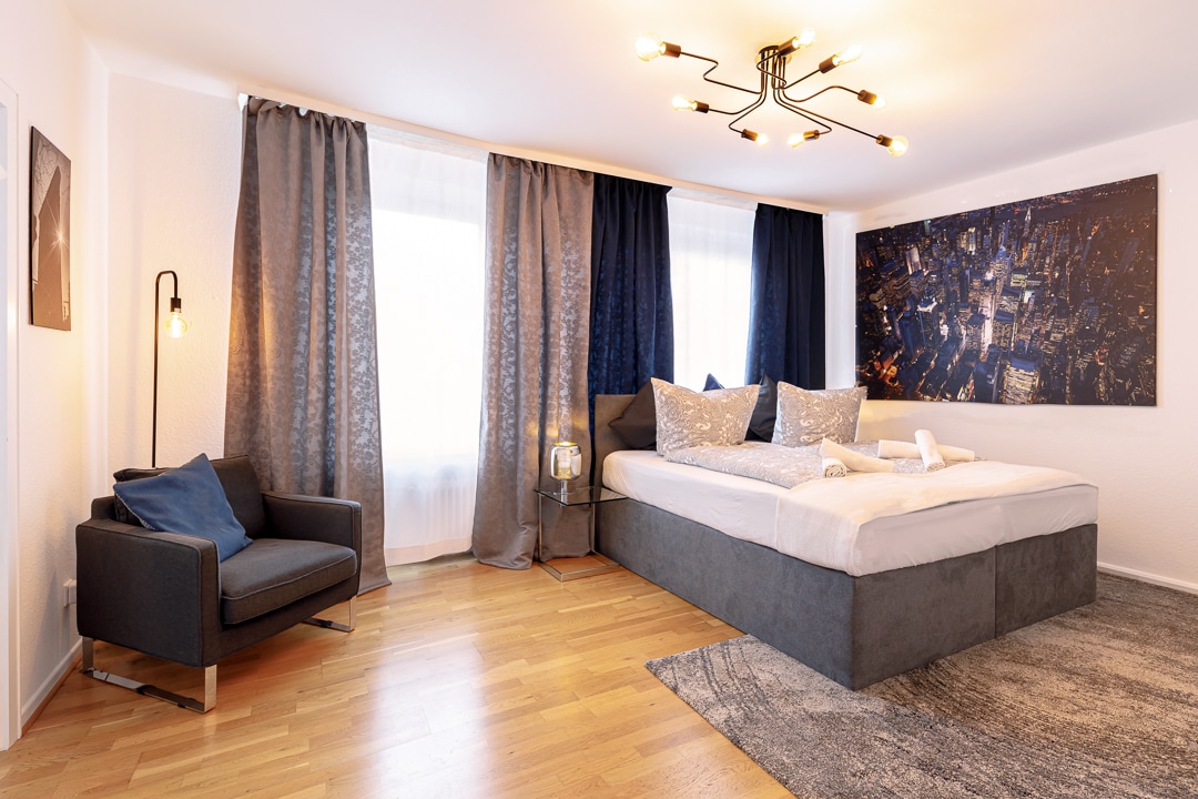 A spacious sleeping area features a large bed adorned with pillows and a decorative throw. A cozy reading corner with a chair is situated nearby. The room is illuminated by warm lighting, and large windows are framed by dark curtains, providing natural light.
