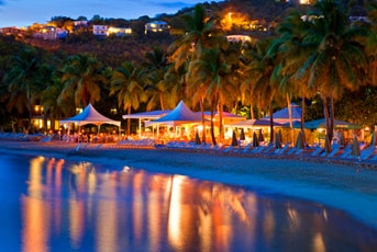 The beachfront area is illuminated by soft lights from the surrounding resort, creating a warm ambiance. Palm trees line the shore, reflecting in the calm water, while lounge chairs and tents provide spaces for relaxation. The distant hillside is dotted with twinkling lights from nearby accommodations.