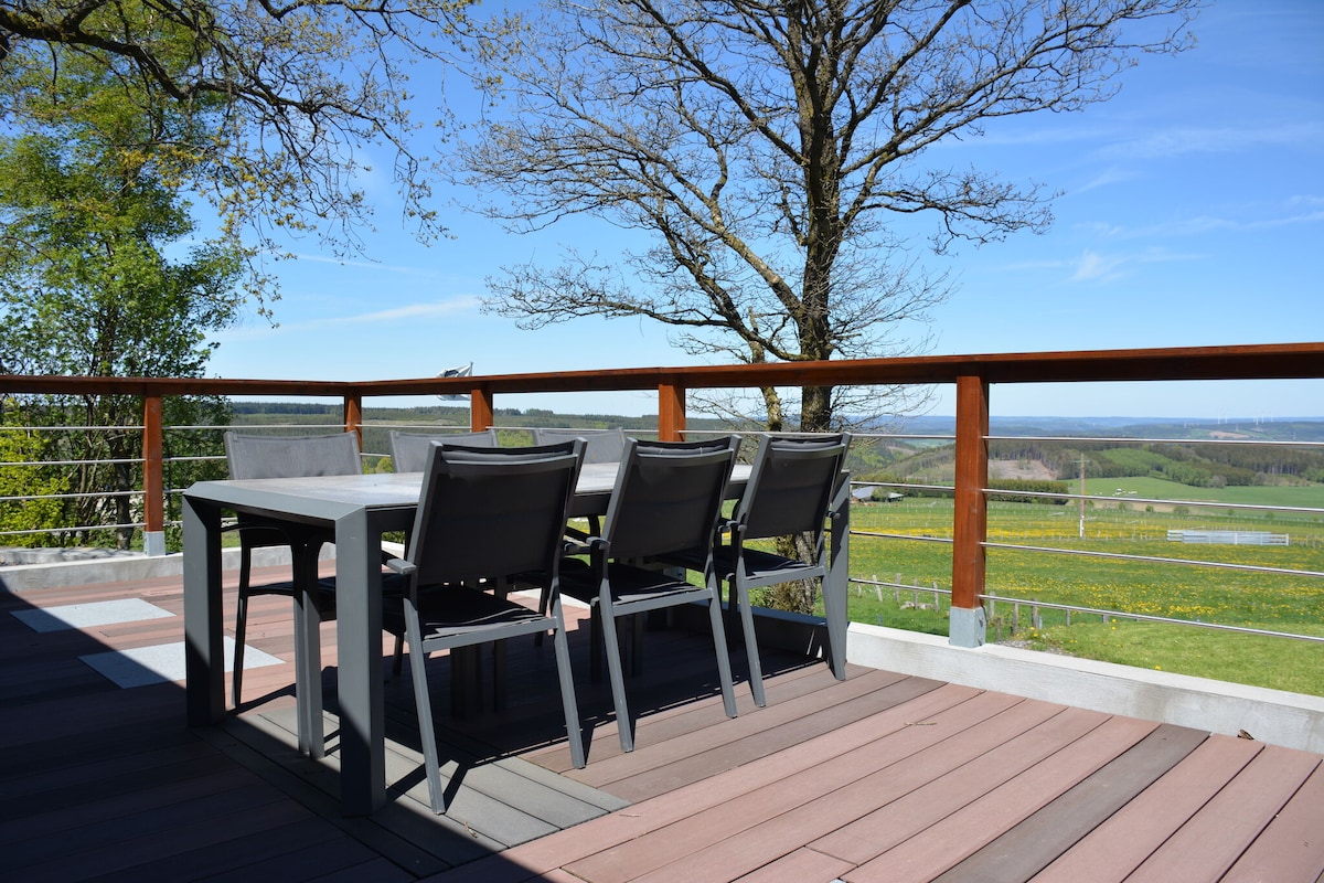A deck area features a rectangular dining table surrounded by six gray chairs. The space offers views of the surrounding valleys and forest, with a wooden railing providing safety and enhancing the outdoor experience.