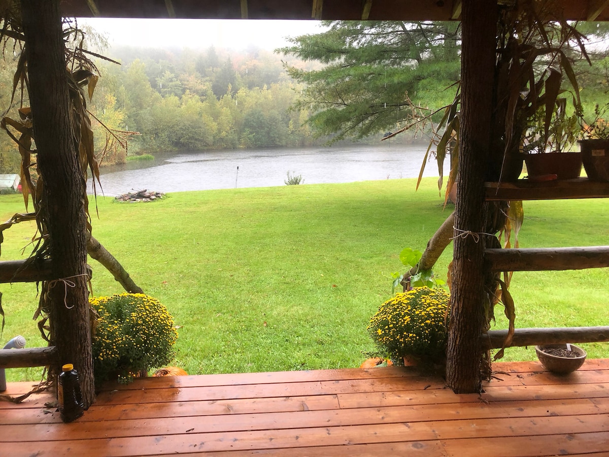 A serene view from the cabin's porch showcases a lush green lawn leading to a calm pond. Decorative flower pots with yellow blooms frame the sides, while misty trees line the background. A firepit is visible on the opposite bank, hinting at outdoor activities.