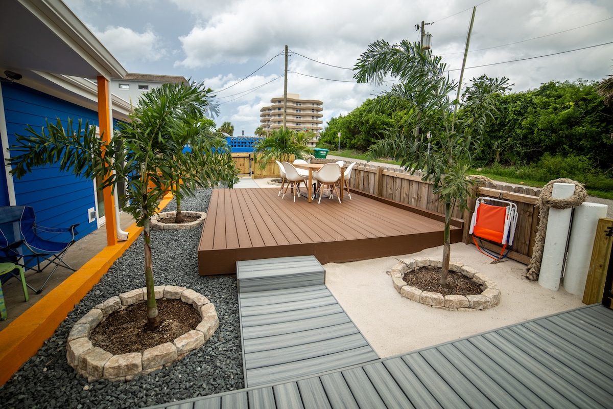 The outdoor area features a large wooden deck surrounded by palm trees and landscaped beds. A dining set with four chairs sits atop the deck, while a charcoal grilling station is positioned nearby. Pathways of grey composite and stones lead through the space.