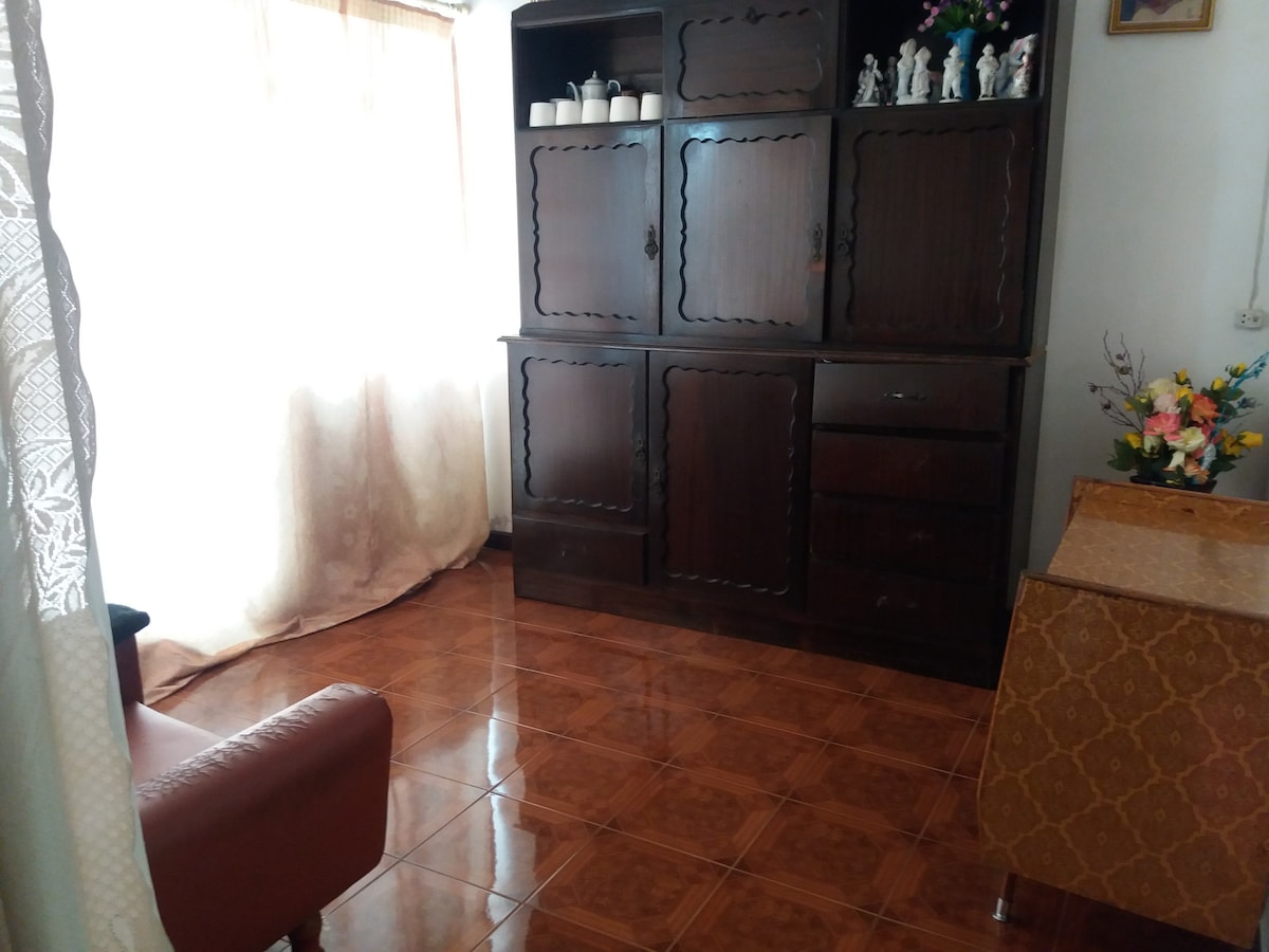 A wooden cabinet with decorative items is positioned against a brightly lit wall, providing storage and display. The space features polished tile flooring that reflects the natural light coming from a large window with light curtains. A cozy armchair adds to the welcoming atmosphere.