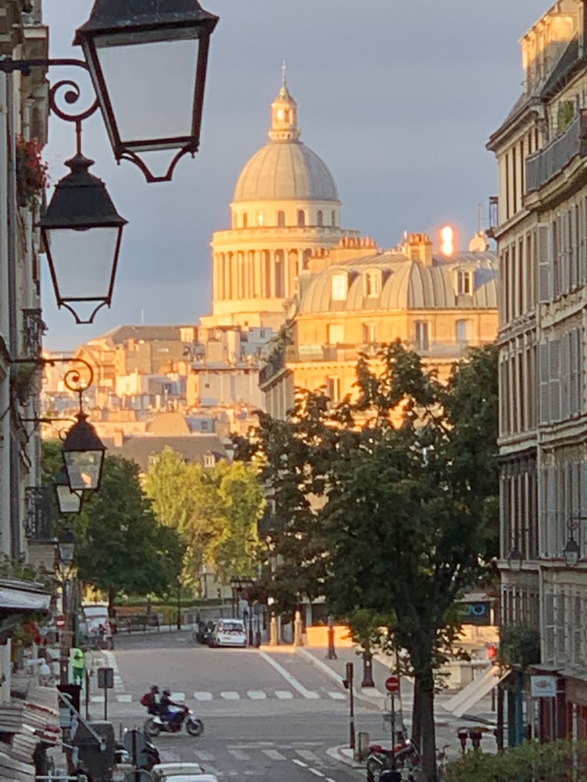 A city street is depicted, leading toward a large dome structure illuminated by soft light. Trees line the avenue, while vintage street lamps are visible in the foreground. The scene captures a calm urban atmosphere with buildings showcasing classic architectural details.