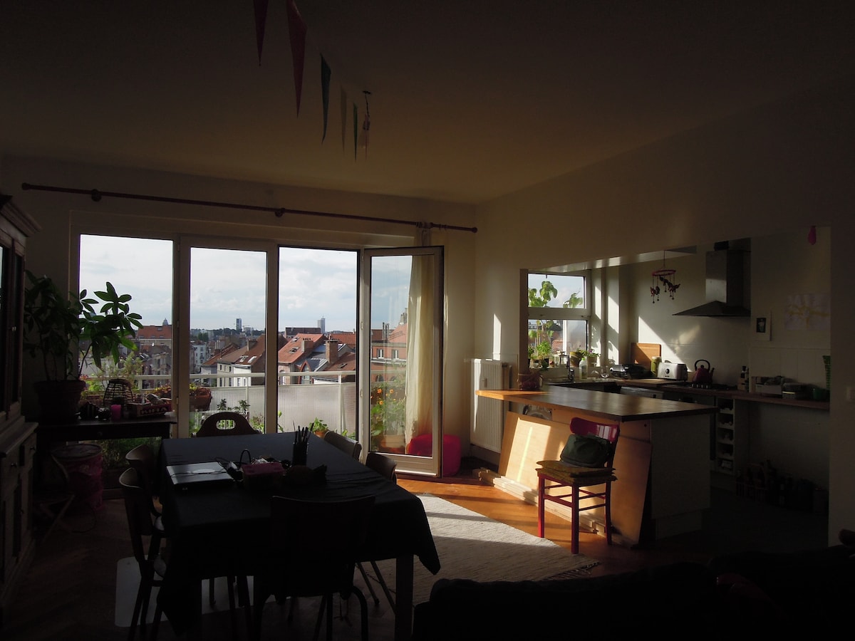A well-lit living area features large glass doors opening to a balcony, providing a view of the surrounding rooftops. A dining table with chairs, and a kitchen equipped with modern appliances, are visible. Light and greenery create an inviting atmosphere.