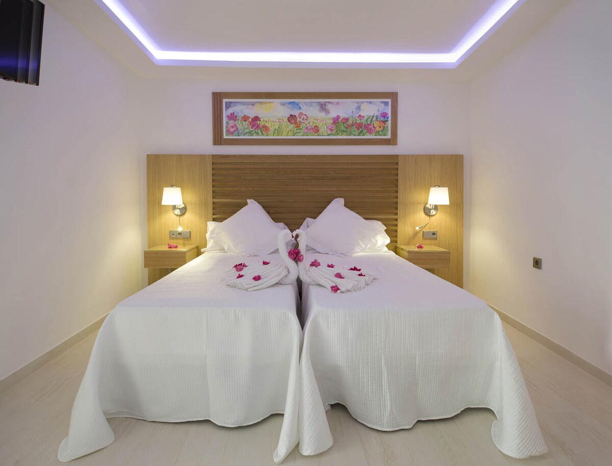 A tranquil sleeping area features two spacious beds dressed in white linens, with bedside lamps providing soft illumination. A decorative wall art piece above the headboard adds a touch of color, and subtle ambient lighting enhances the overall warmth of the space.